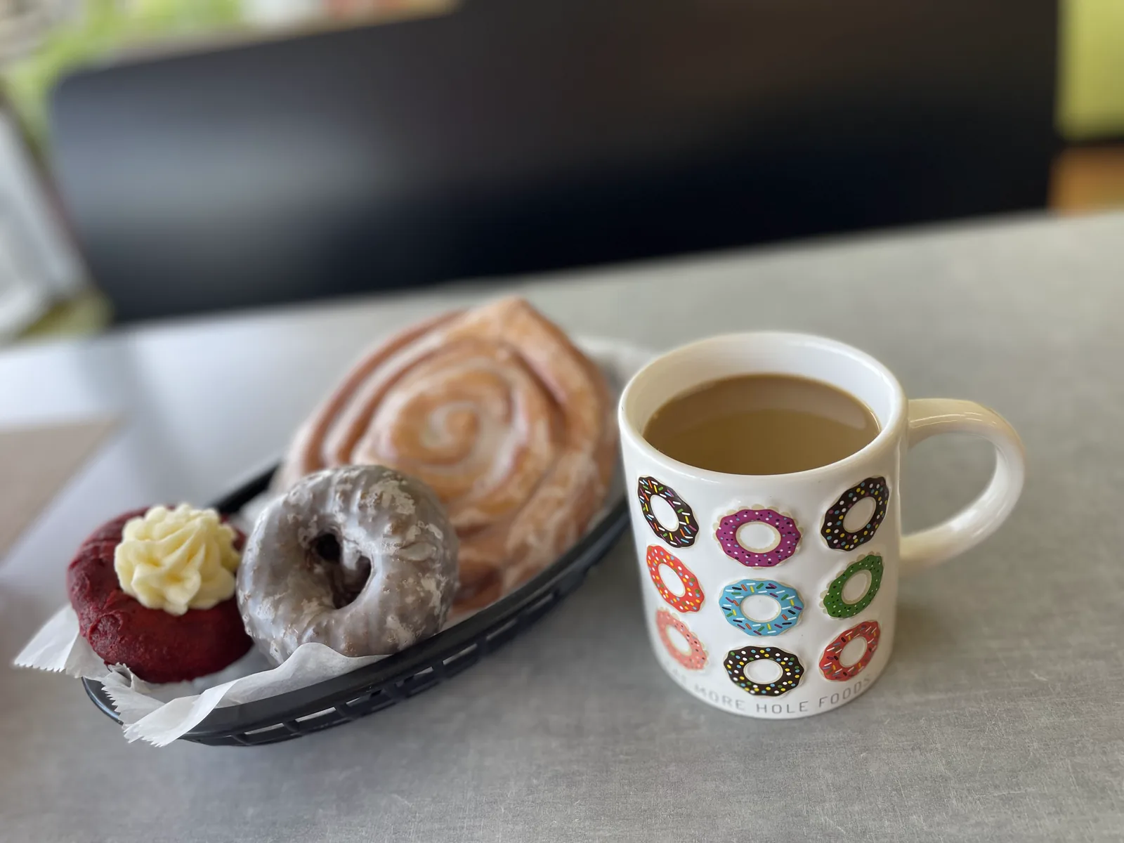 A mug of hot coffee beside a basket holding a cinnamon roll, a glazed donut and a red velvet donut