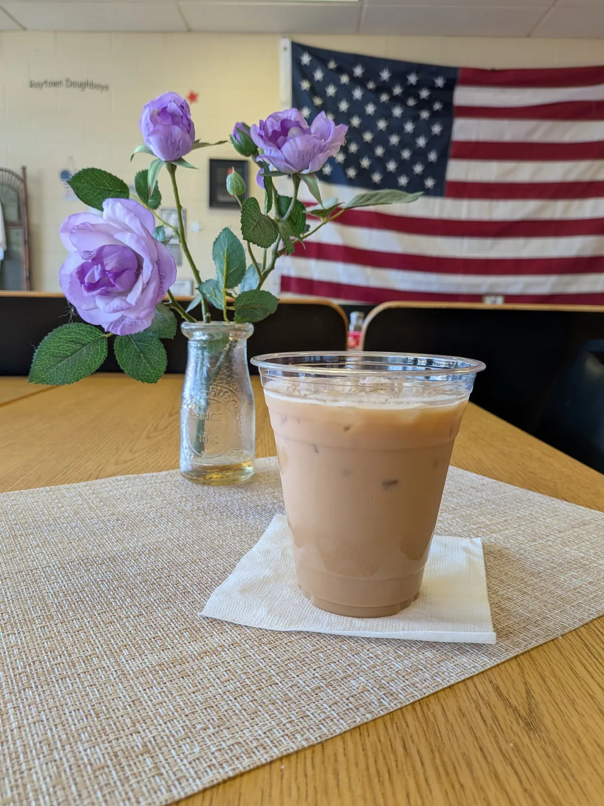 An iced latte on a table in the Doughboys dining room, with flowers and the flag behind it