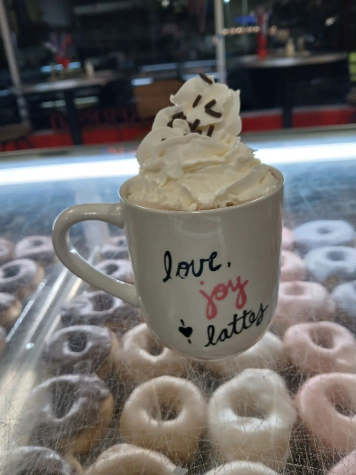 A whipped cream topped latte held above a tray of freshly glazed donuts